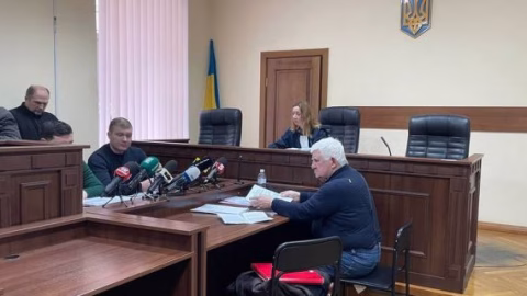 Ukrainian military courtroom scene with detained generals escorted by officers, press and courthouse backdrop representing war accountability.